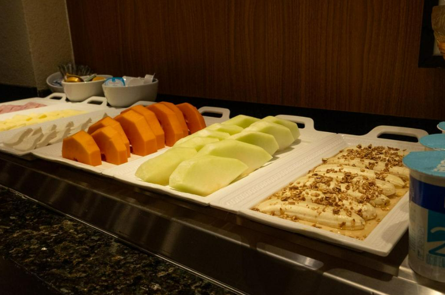 Frutas e acompanhamentos em uma bandeja branca de buffet: mamão, melão, bananas com nozes, iogurte.