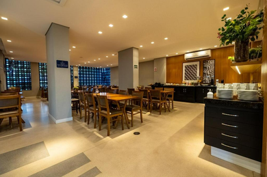 Área de café da manhã do hotel com mesas, cadeiras, bufê e grandes janelas.