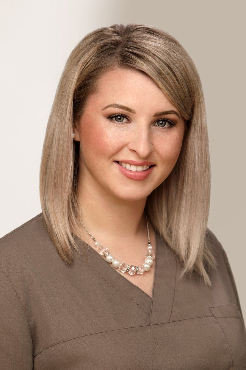 Woman with blonde hair, smiling, wearing brown top and pearl necklace.