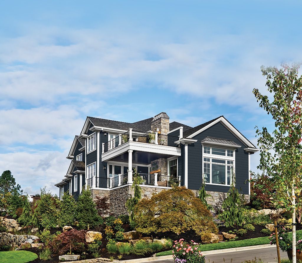 A multi-level dark gray house with a stone base, large windows, and white trim, nestled in a lush, green landscape.