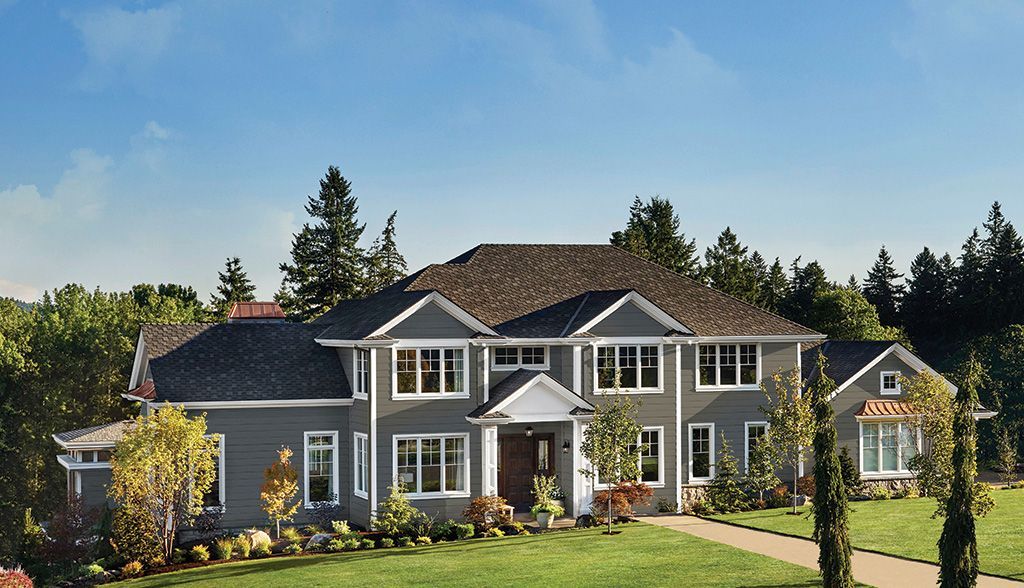 A large, two-story grey house with a dark shingled roof, set against a backdrop of trees under a clear blue sky.