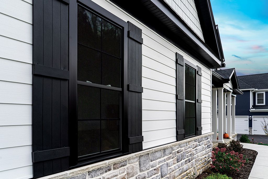 Close-up of a house exterior featuring white horizontal siding, black window shutters, and a stone foundation.