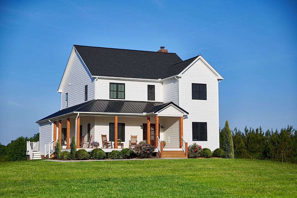 A two-story white farmhouse with a black metal roof, wrap-around porch, and wooden posts, set in a green grassy field.
