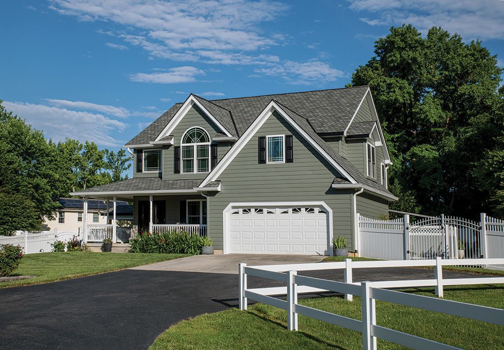 A two-story, sage green house with a white garage door, a wrap-around porch, and a white picket fence on a sunny day.