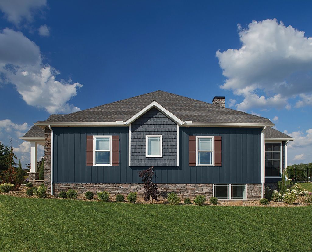 A blue house with vertical siding, brown shutters, and stone foundation under a blue sky with white clouds.