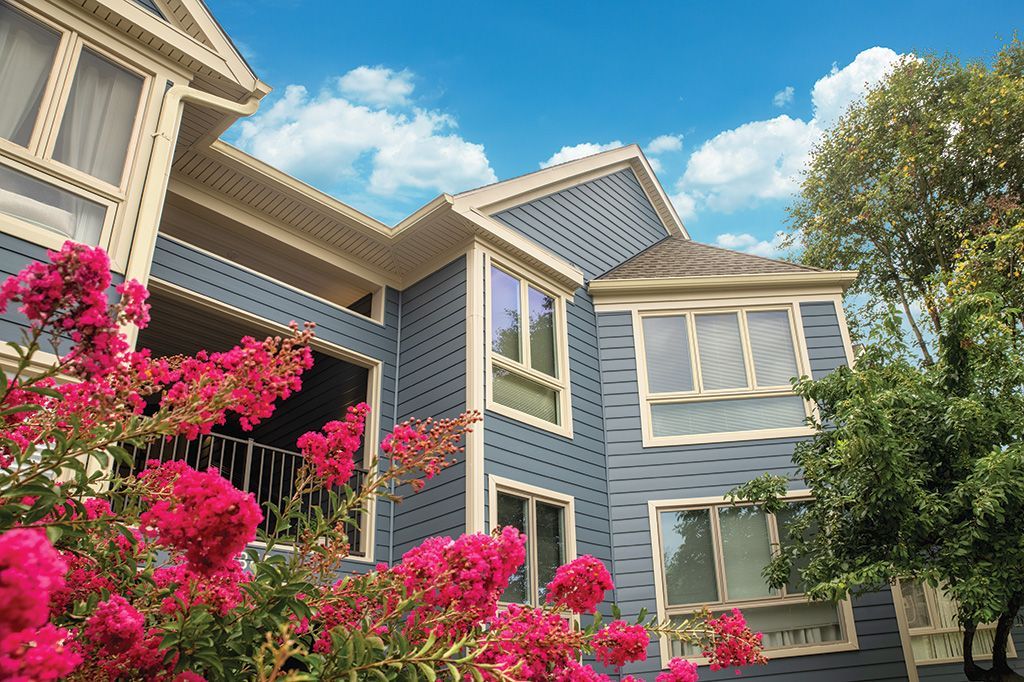 Blue multi-story house with white trim, framed by bright pink crape myrtle flowers under a clear blue sky.
