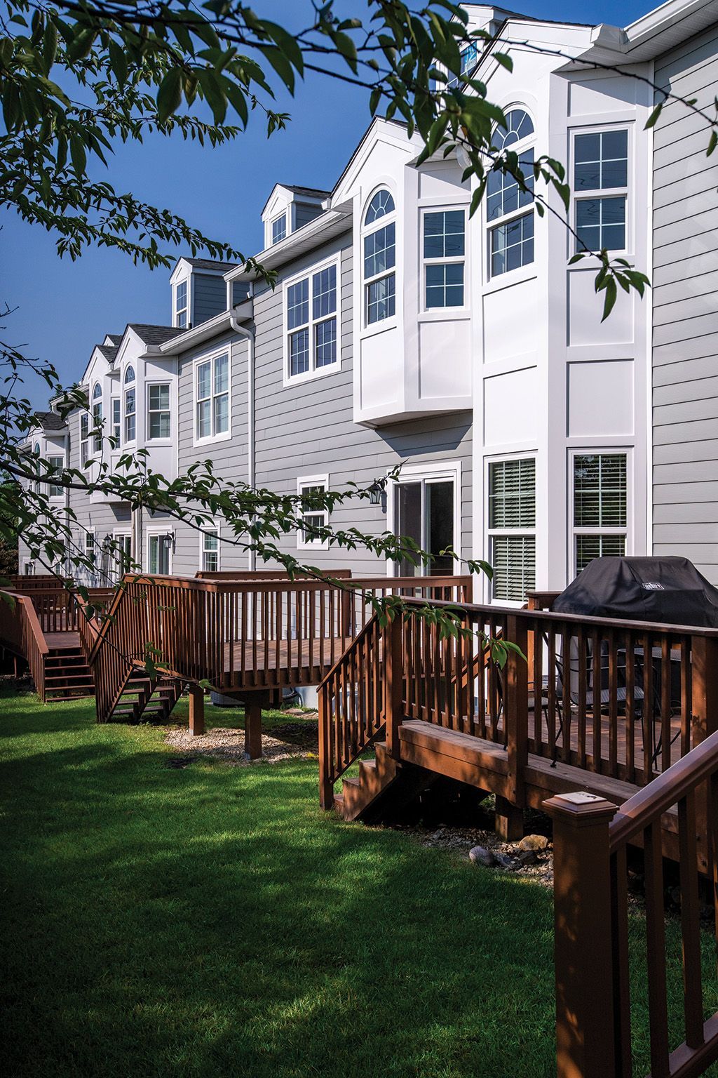 A row of gray townhouses with wooden decks extending into a green, grassy yard on a sunny day.