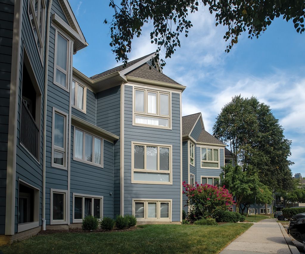 A blue, multi-story apartment complex with white trim and multi-pane windows sits beside a sidewalk and grassy lawn.