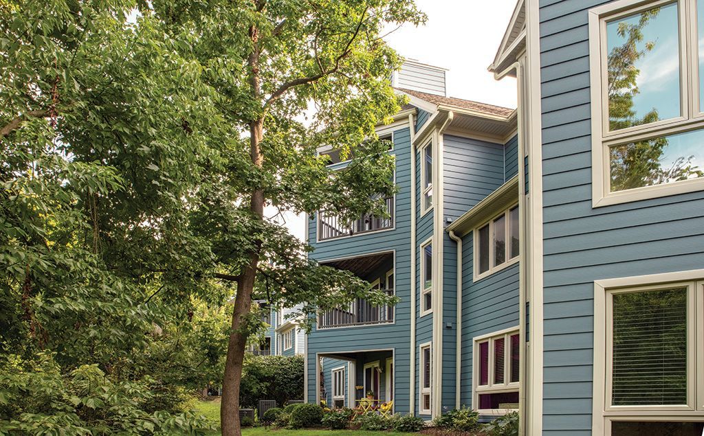 A multi-story blue building with white trim set among lush green trees and garden beds.