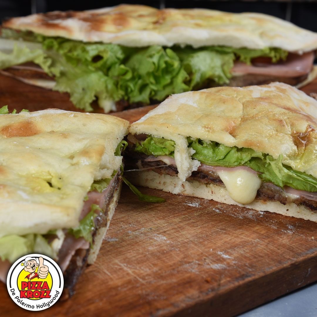 Dos mitades de un sándwich de pan plano con queso derretido, jamón y lechuga fresca, sobre una tabla de cortar de madera.