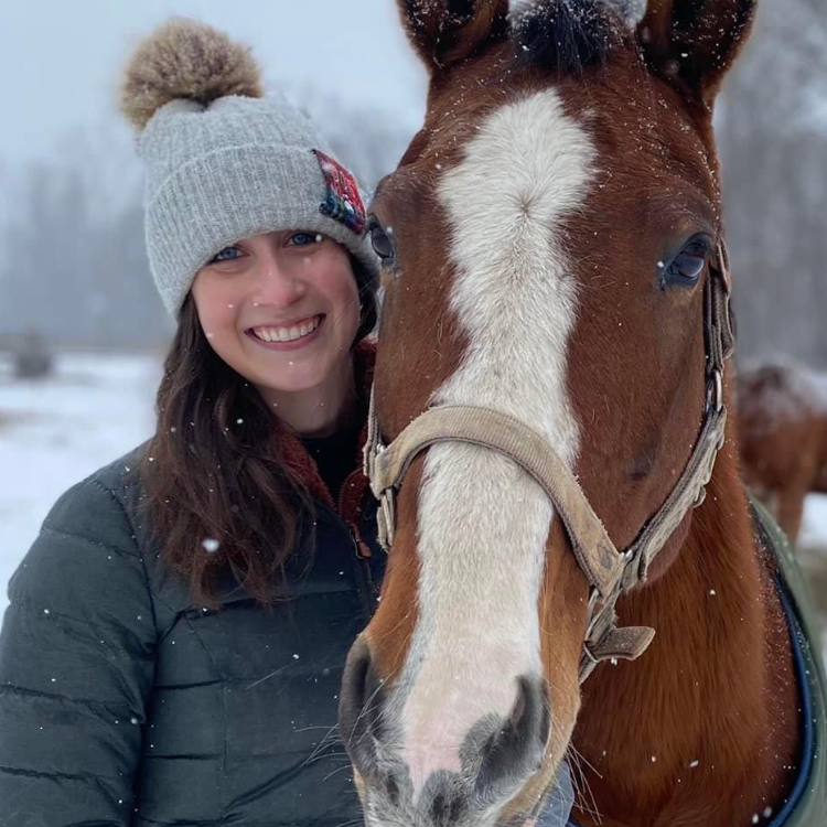 A woman in a gray hat is standing next to a brown horse in the snow.