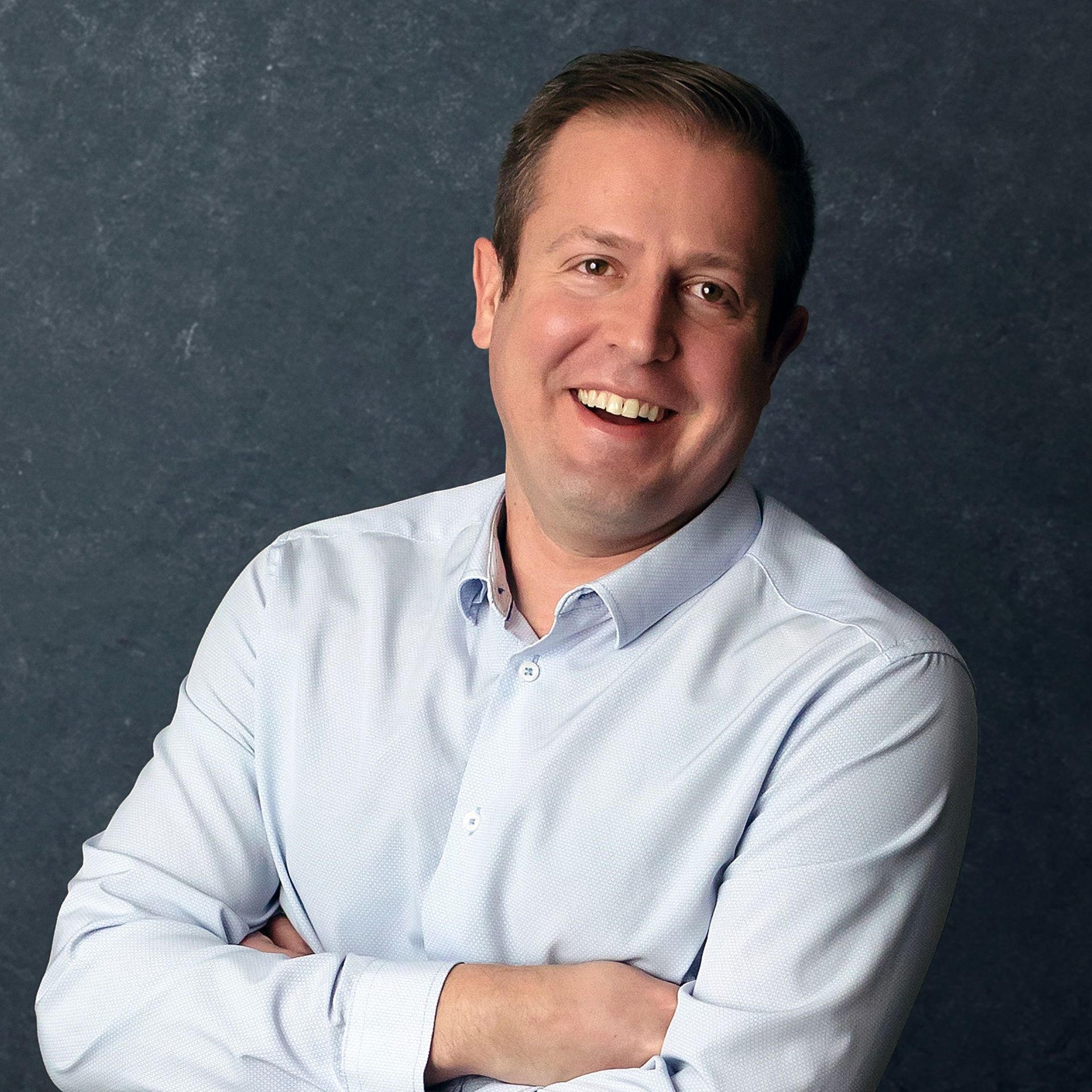 a man in a light blue shirt smiles with his arms crossed