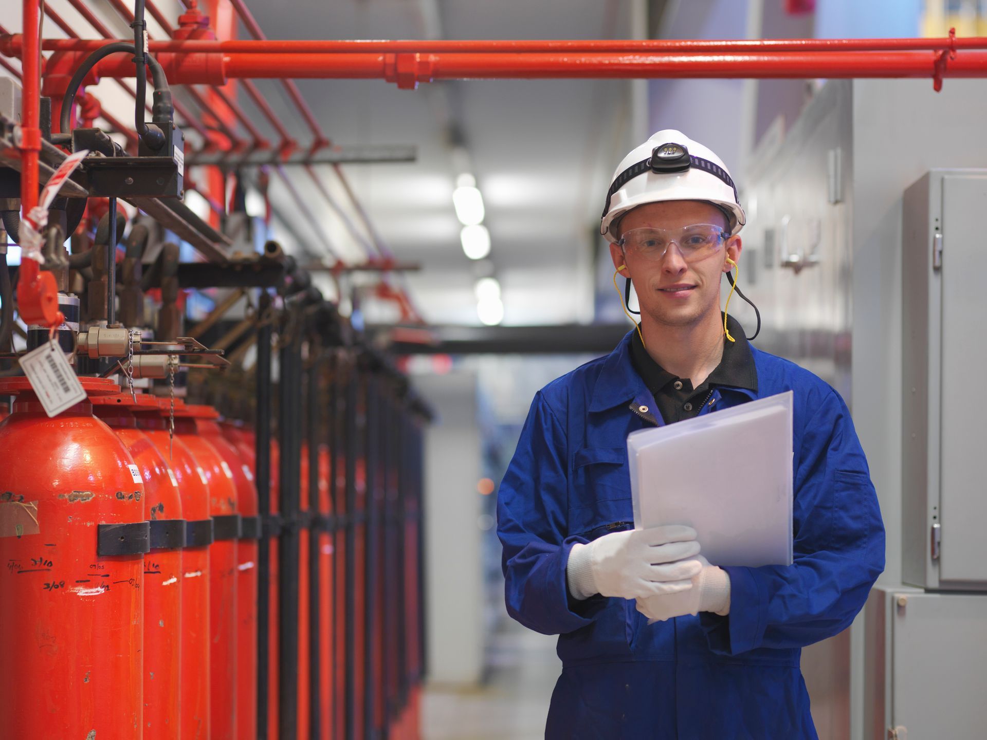 Male engineer wears blue workwear inside silver turbine hall, next to red tanks.