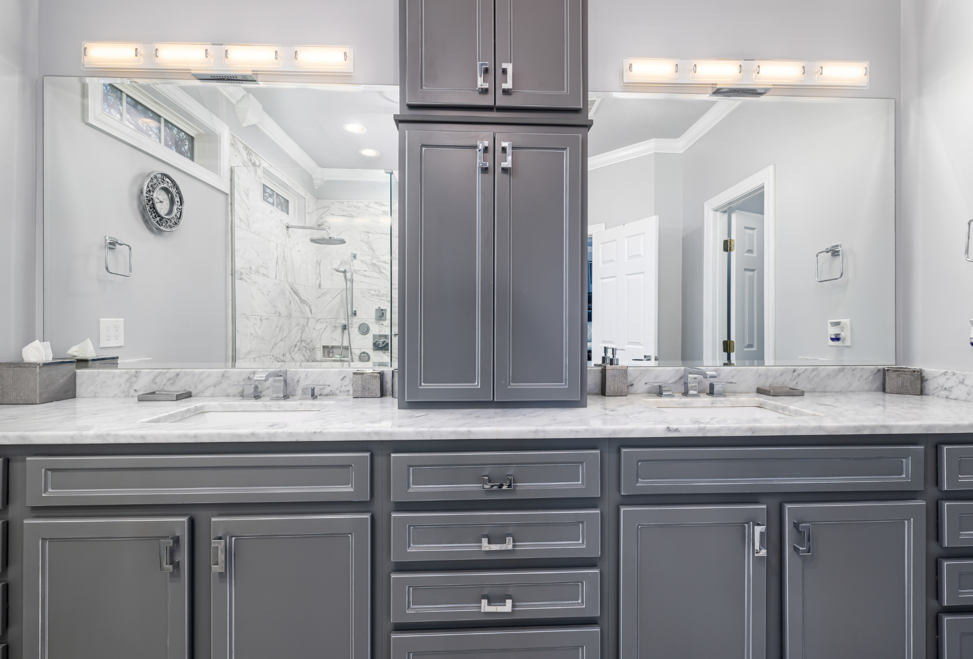 Gray bathroom vanity with two sinks, two large mirrors, and a tall cabinet. Light fixtures above the mirrors.