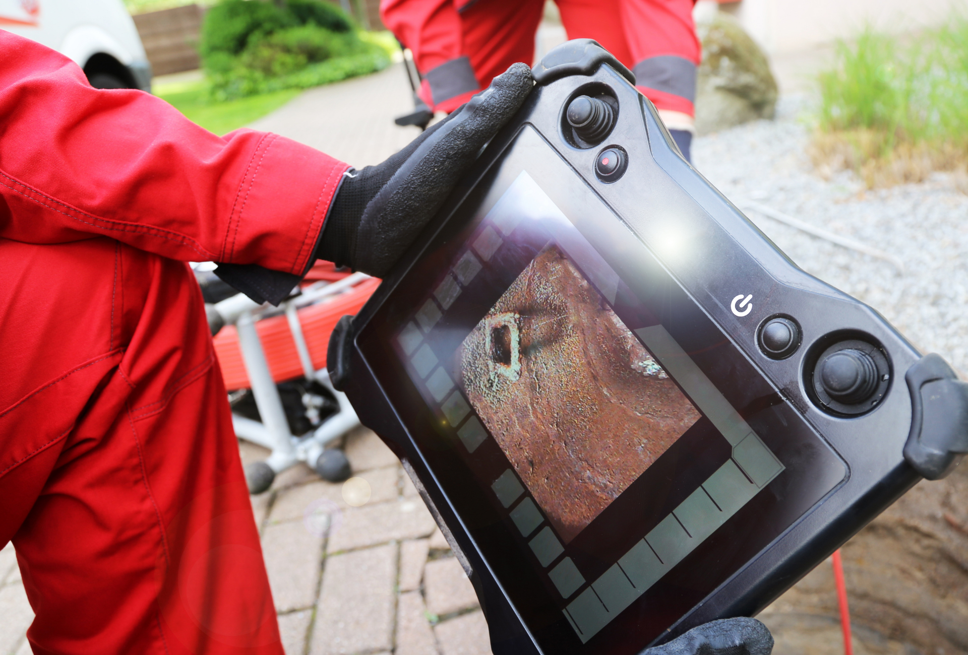 Person in red uniform holding a tablet displaying a close-up of a hole, possibly for inspection, outdoors.