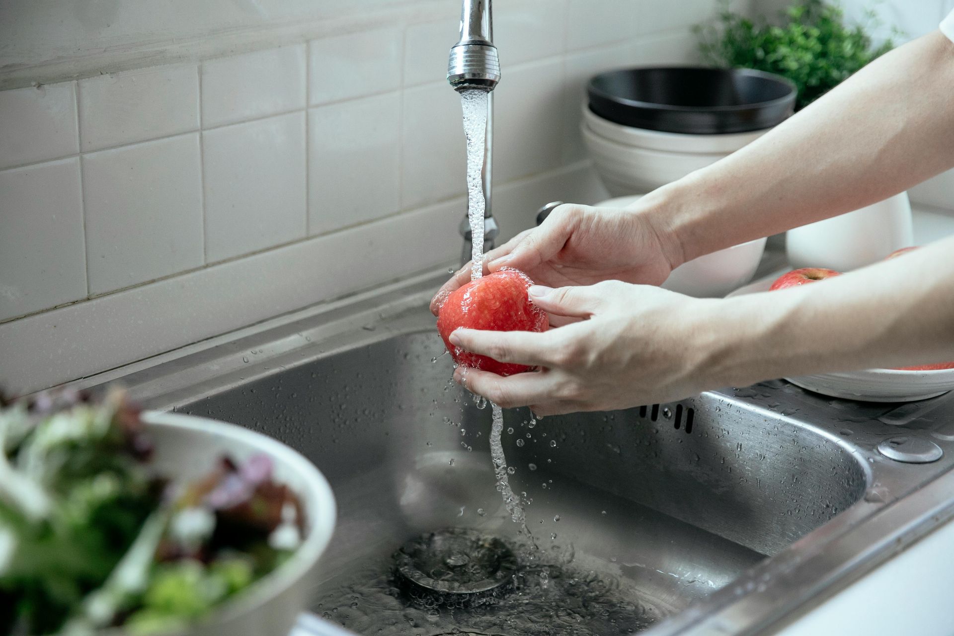 Person washing a red tomato under running water in a stainless steel kitchen sink. Salad bowl is in the foreground.