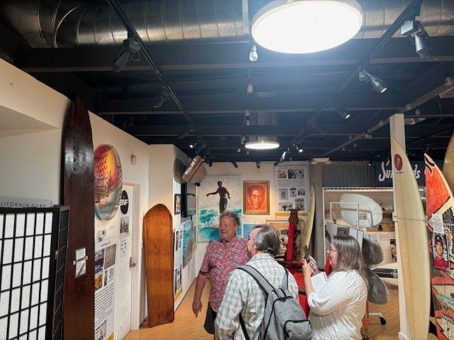 People viewing surfboards in a museum. Wooden, old and modern surfboards on display.