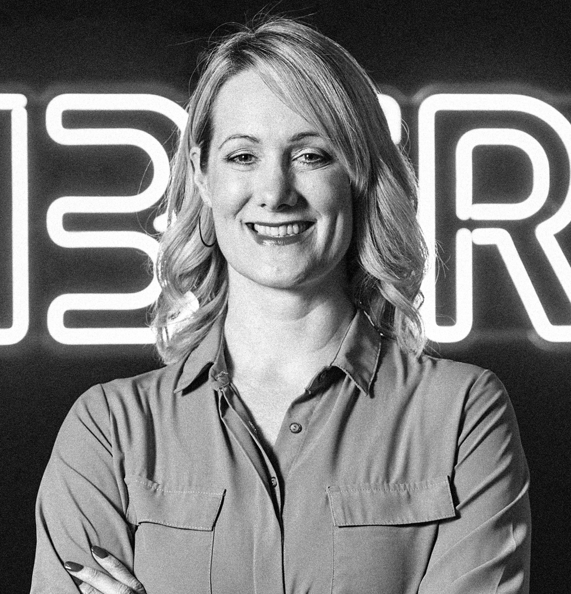 Woman smiling, arms crossed, in front of a neon sign.