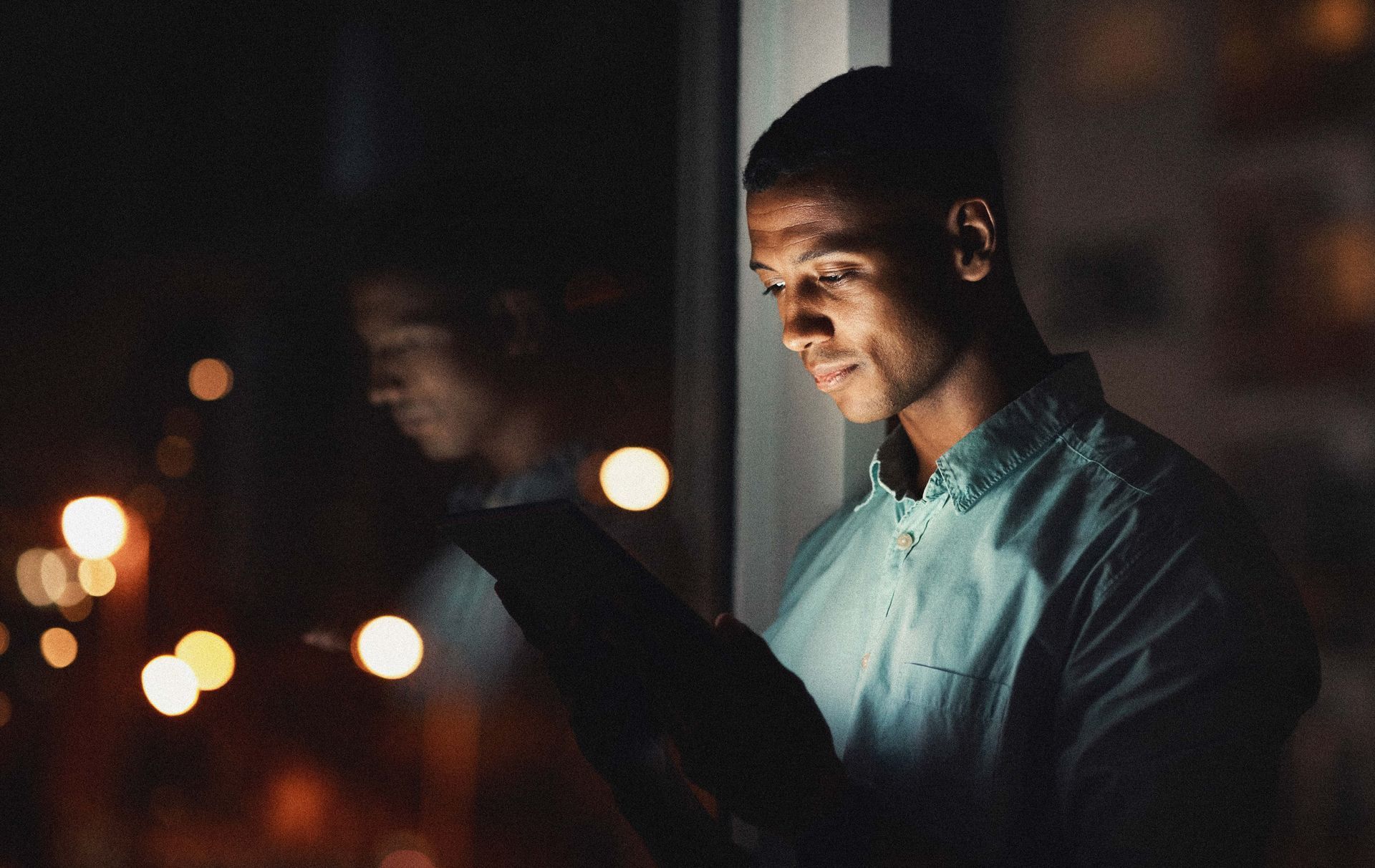 Man looking at tablet near a window at night. City lights reflect in the window.