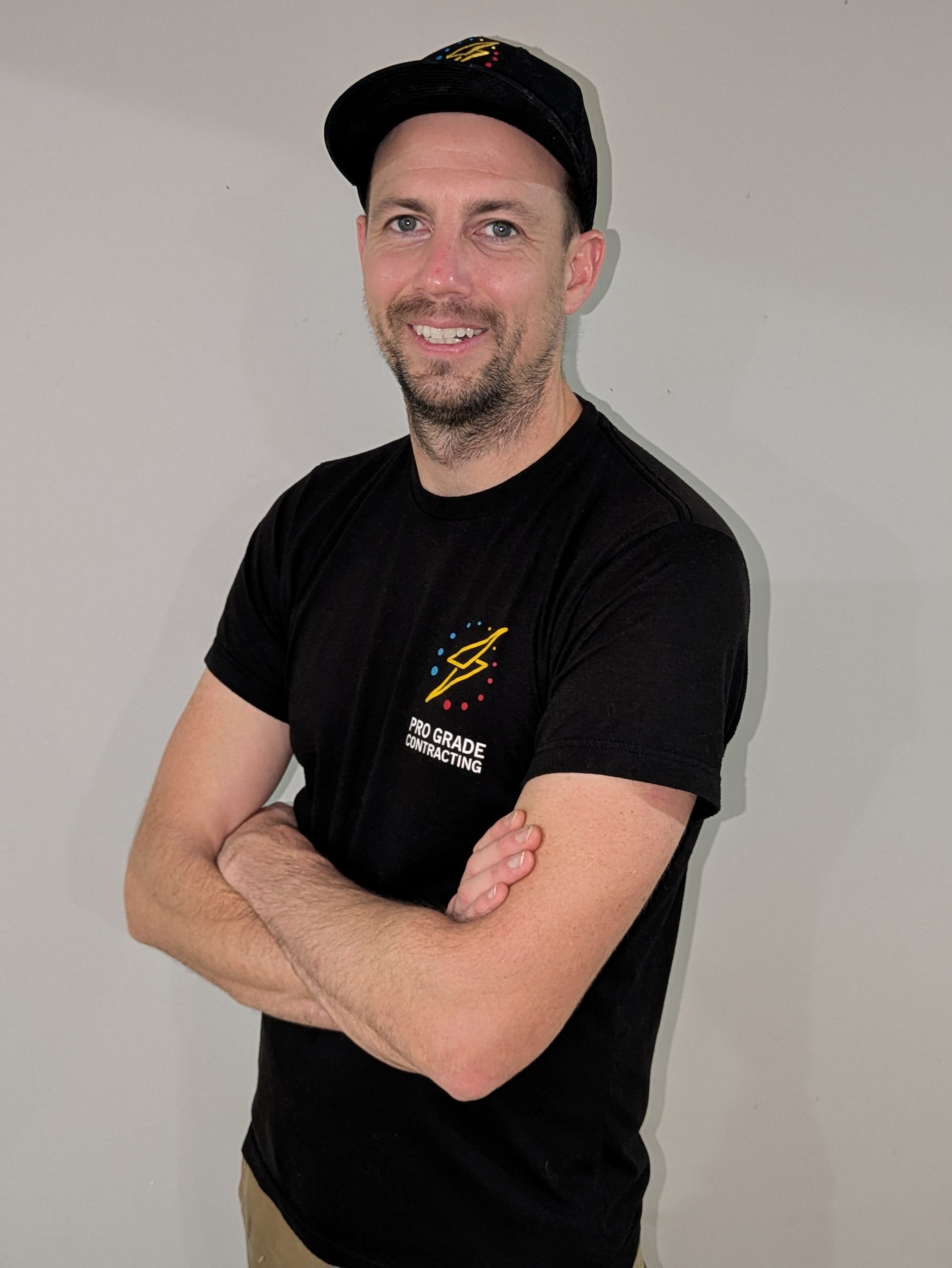 Man in black t-shirt and cap smiles, arms crossed. He stands against a white wall.