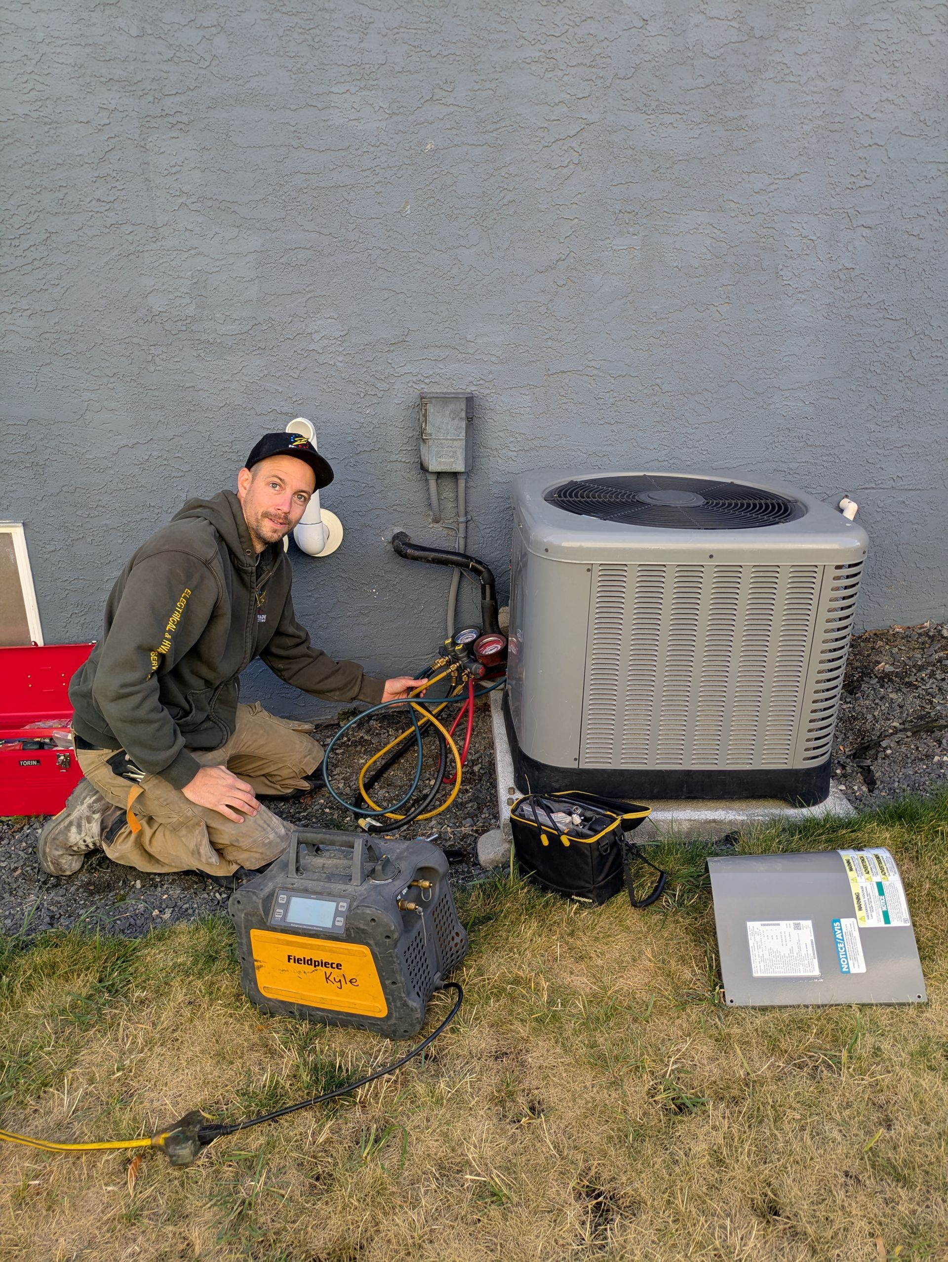 Man kneels near an AC unit, connecting hoses. He's outside on the grass near a wall.