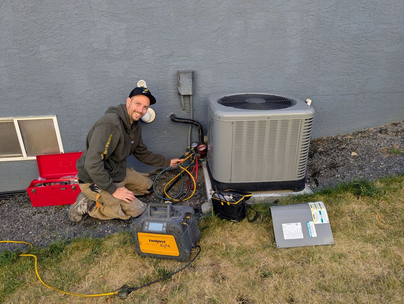 HVAC technician kneeling beside an air conditioning unit; he's working on it with tools and equipment outdoors.