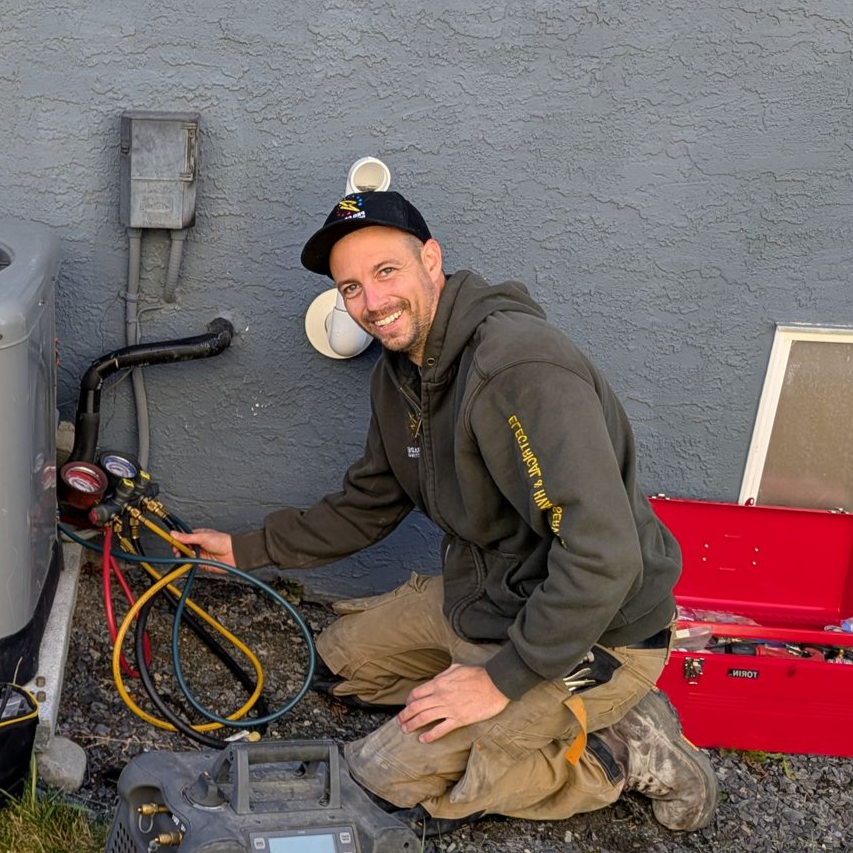HVAC technician kneeling, working on AC unit outside, holding gauges, smiling, wearing hat and work clothes.