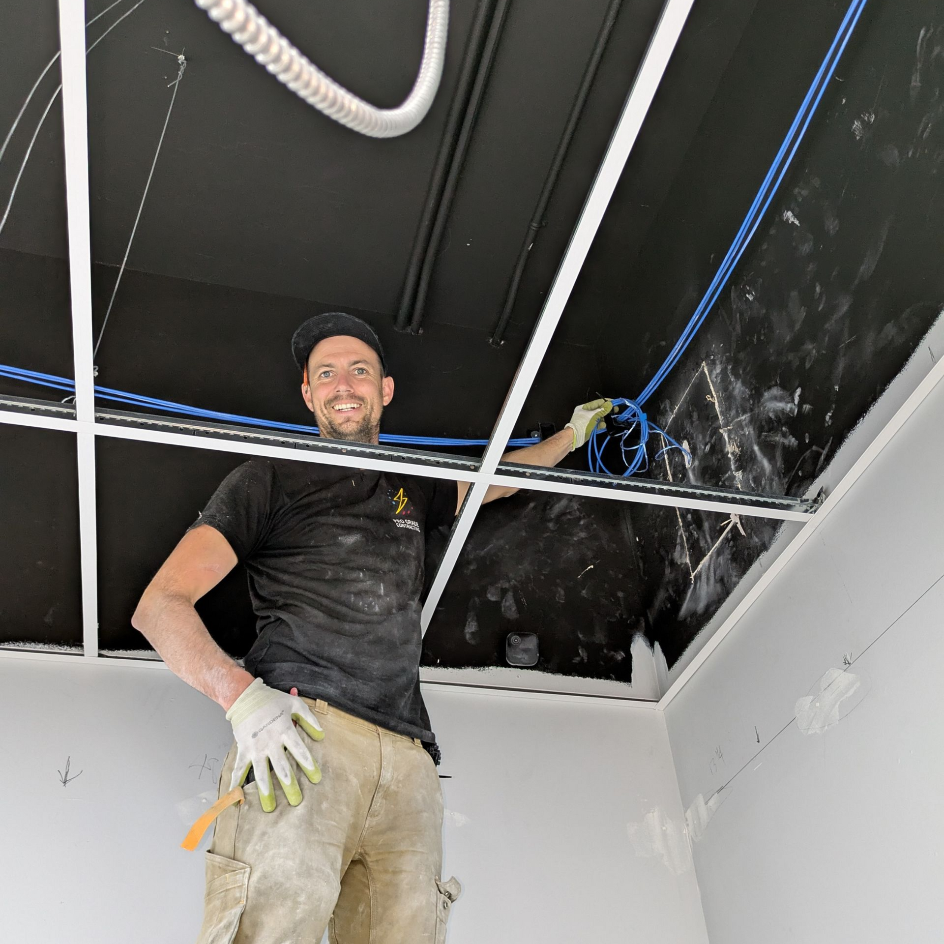 Construction worker installing wiring in a ceiling, wearing gloves and a cap.