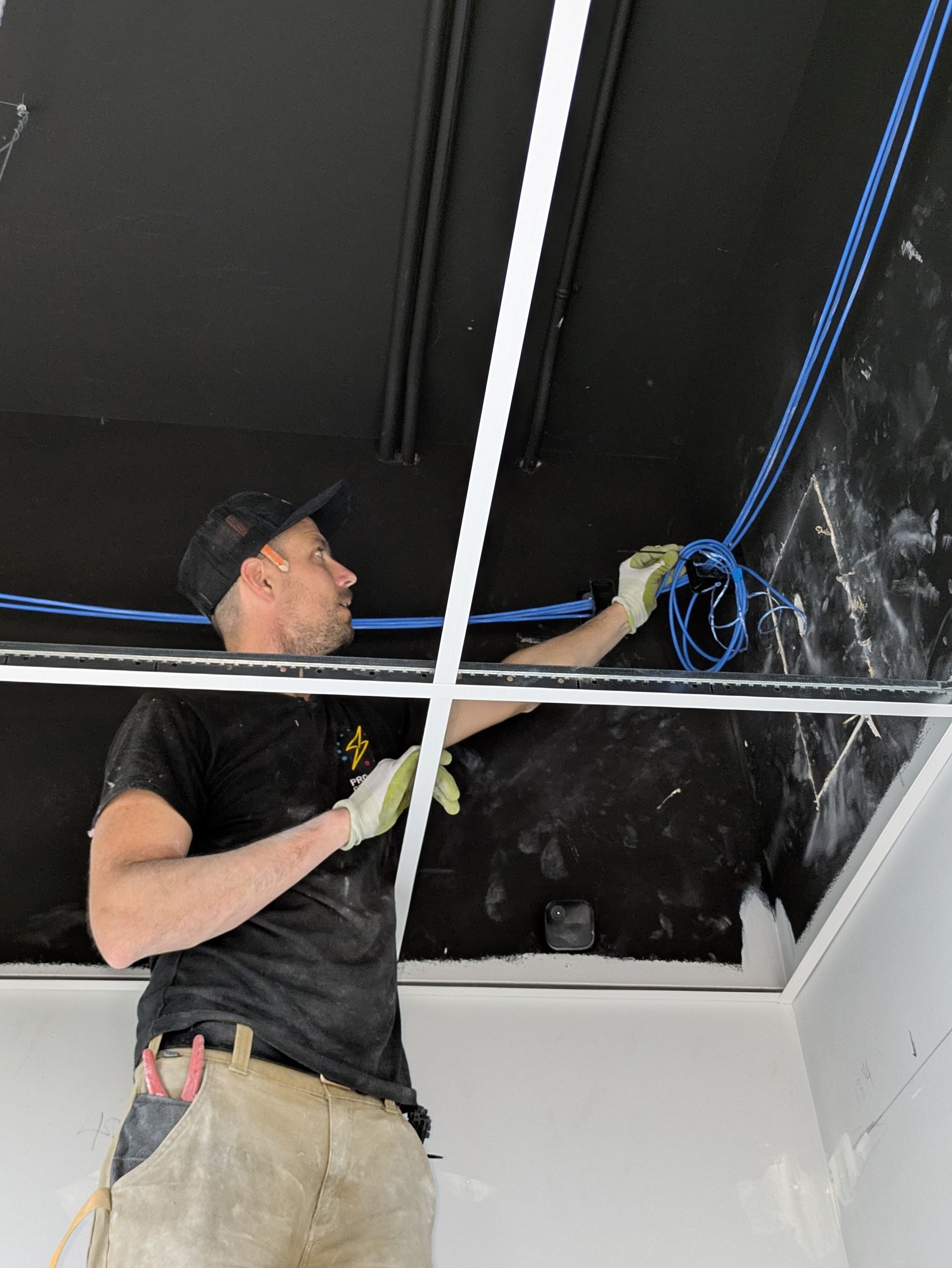 Man in a black shirt installing blue wires on a black ceiling with a white trim.
