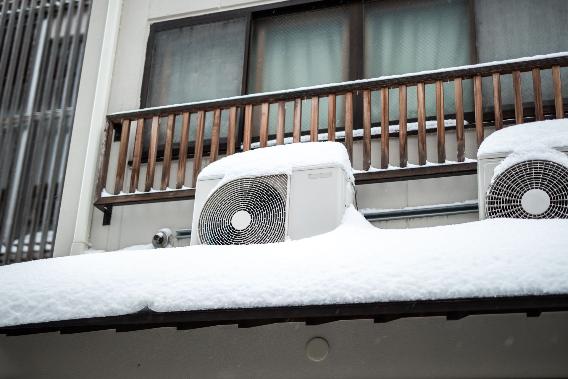 Snow-covered air conditioning units on a rooftop with a wooden balcony and a frosted window.