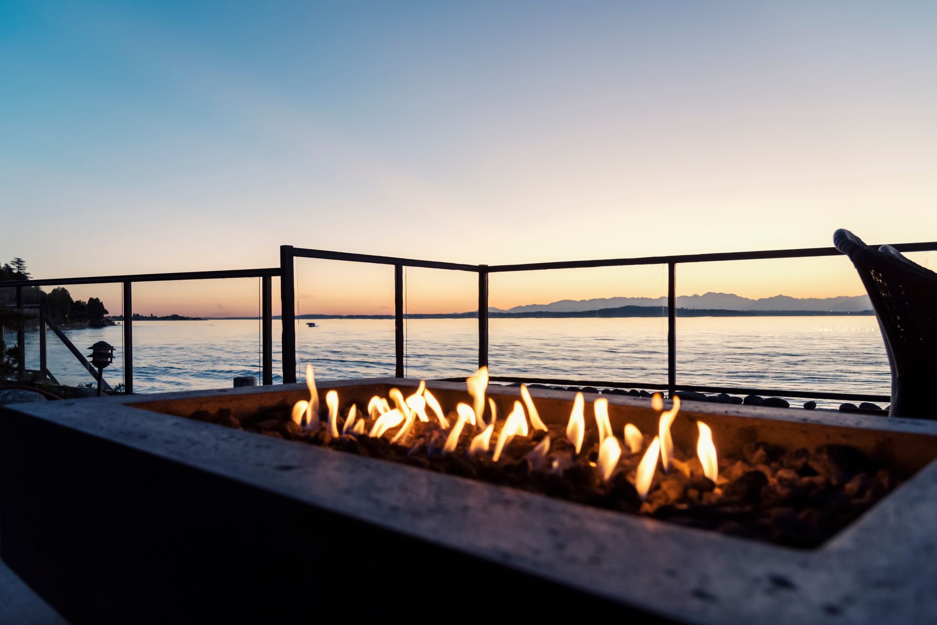 Fire pit flames glow on a waterside patio at dusk.