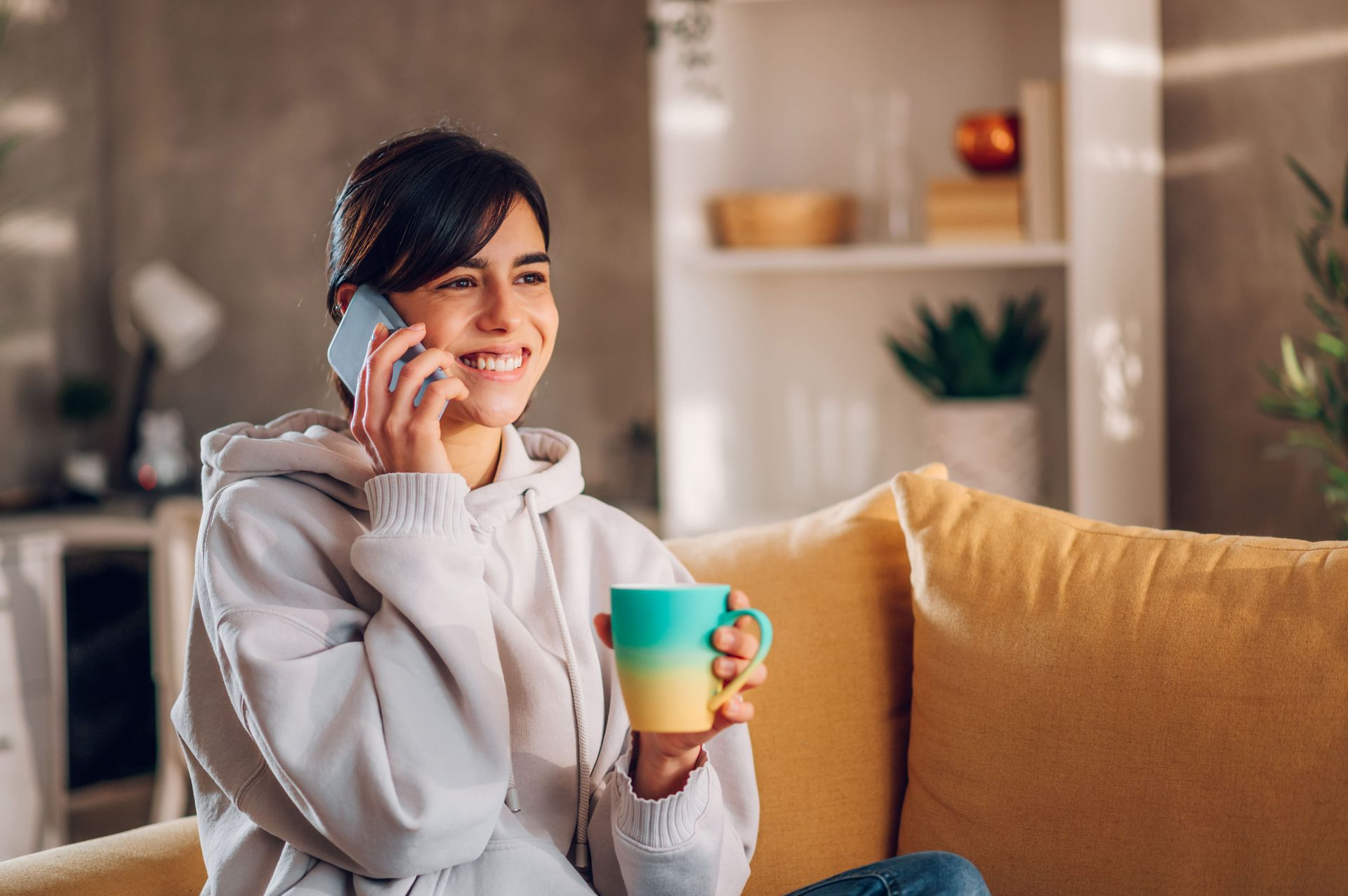 Woman smiling while talking on phone, holding a mug, sitting on a yellow sofa in a well-lit living room.