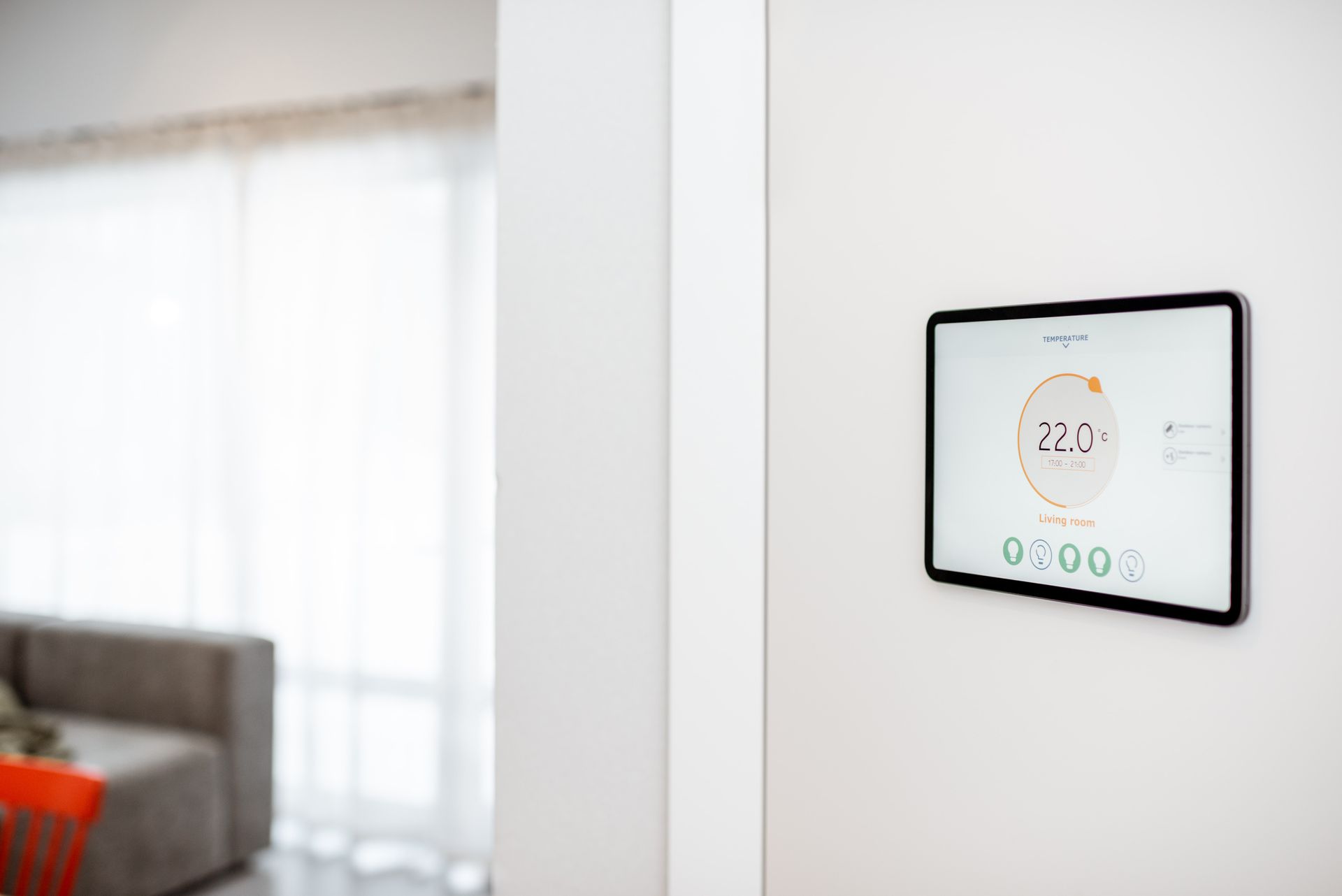 Smart thermostat on a white wall displays 72 degrees, with a living room visible in the background.