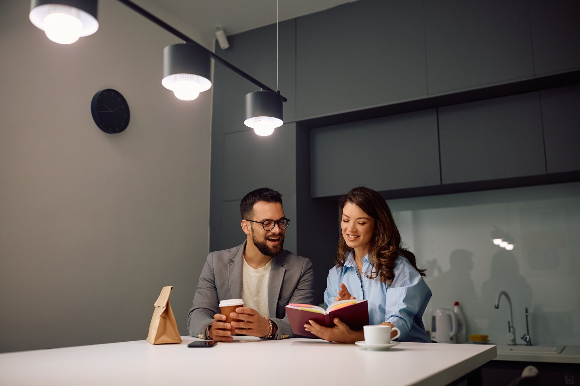 Couple at a kitchen counter, man holding coffee, woman reading a book.