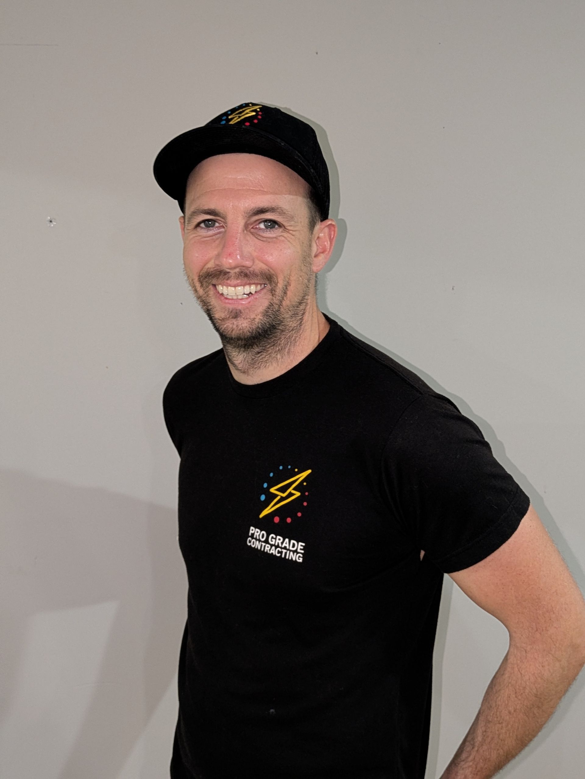 Man in black cap and t-shirt smiles, standing in front of a gray wall.