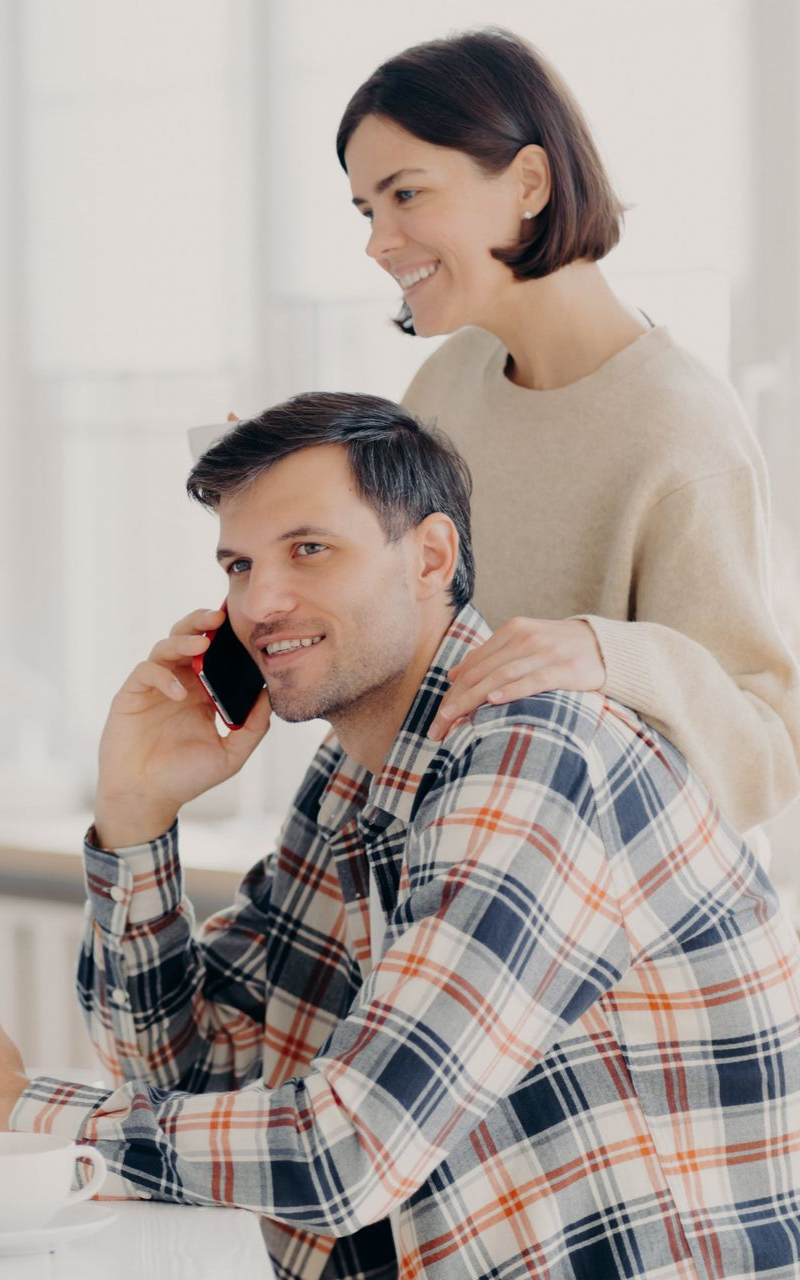 Man on phone, woman smiling behind him, hand on his shoulder. Light setting, plaid shirt.