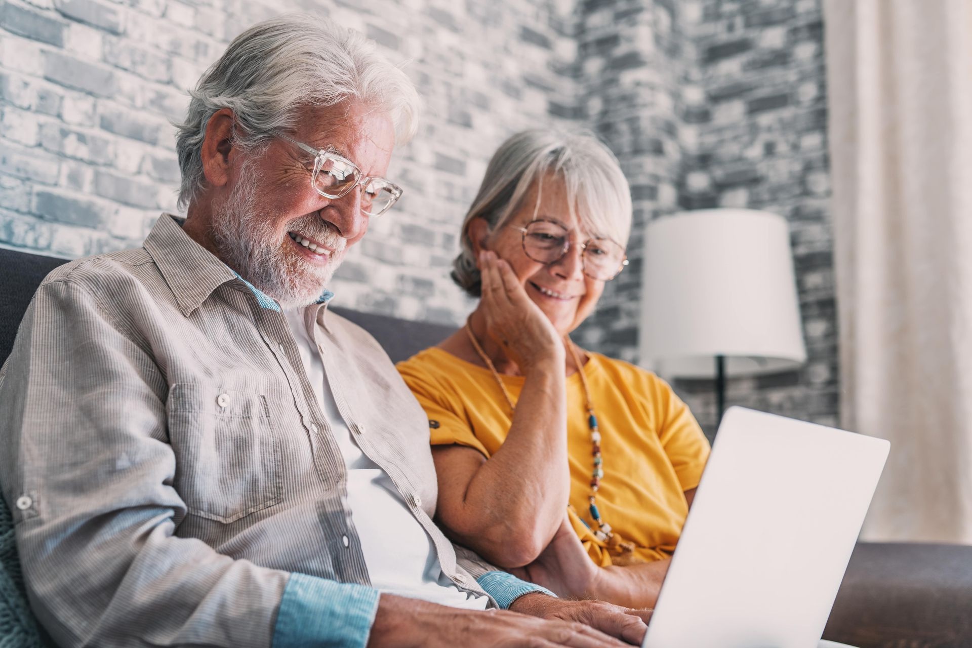 Elderly couple smiling while looking at a laptop computer. Indoors, seated on a couch.