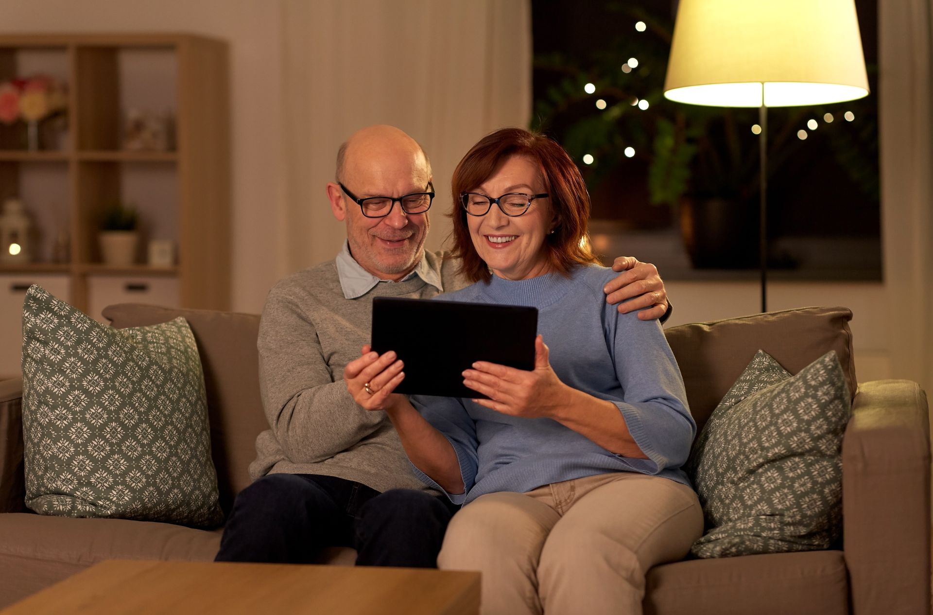 Older couple on couch looking at tablet, smiling. Living room setting, dim lighting.