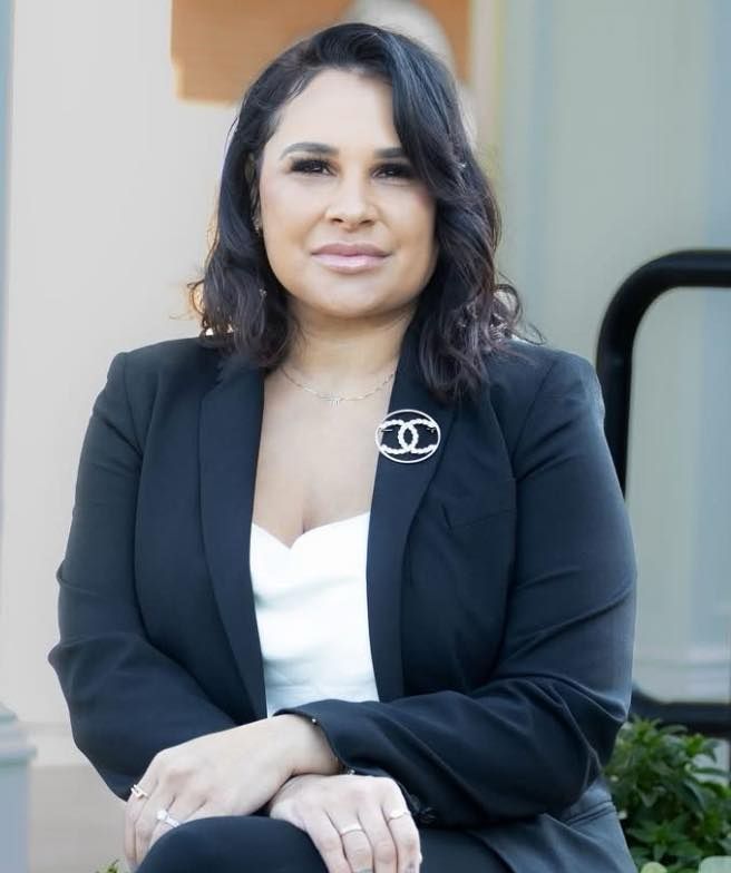 A woman in a black jacket and white top is sitting on a bench.