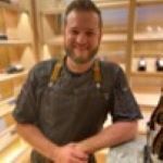 A man with a beard wearing an apron is standing in a kitchen.