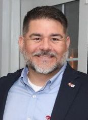 A man with a beard is wearing a blue shirt and a black jacket.