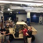 A group of people are standing around a jeep in a room.