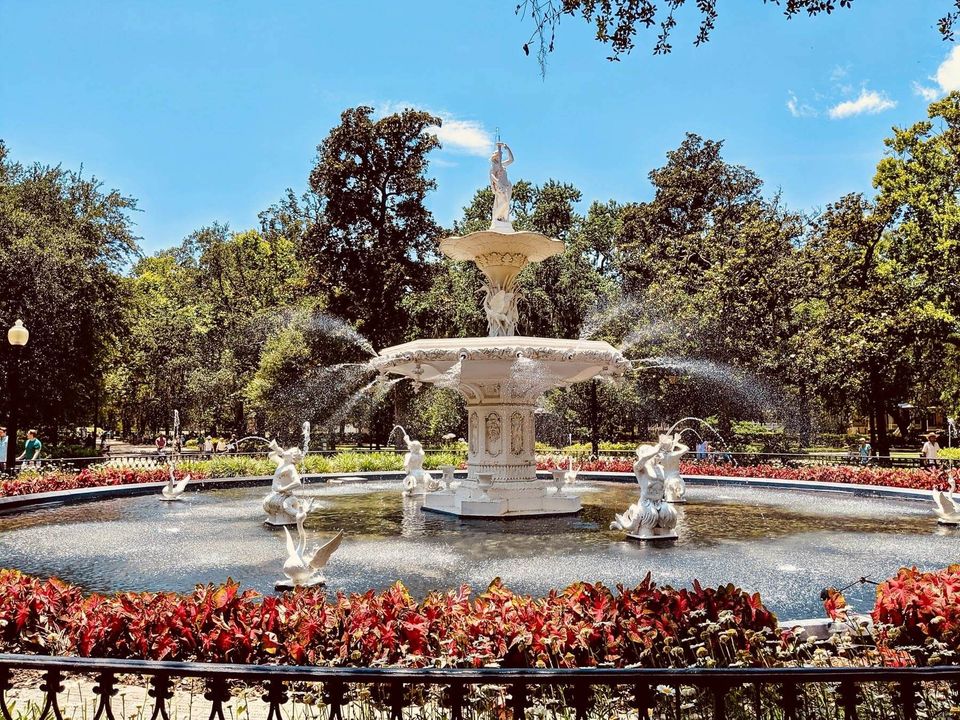 A fountain in a park surrounded by flowers and trees on a sunny day.