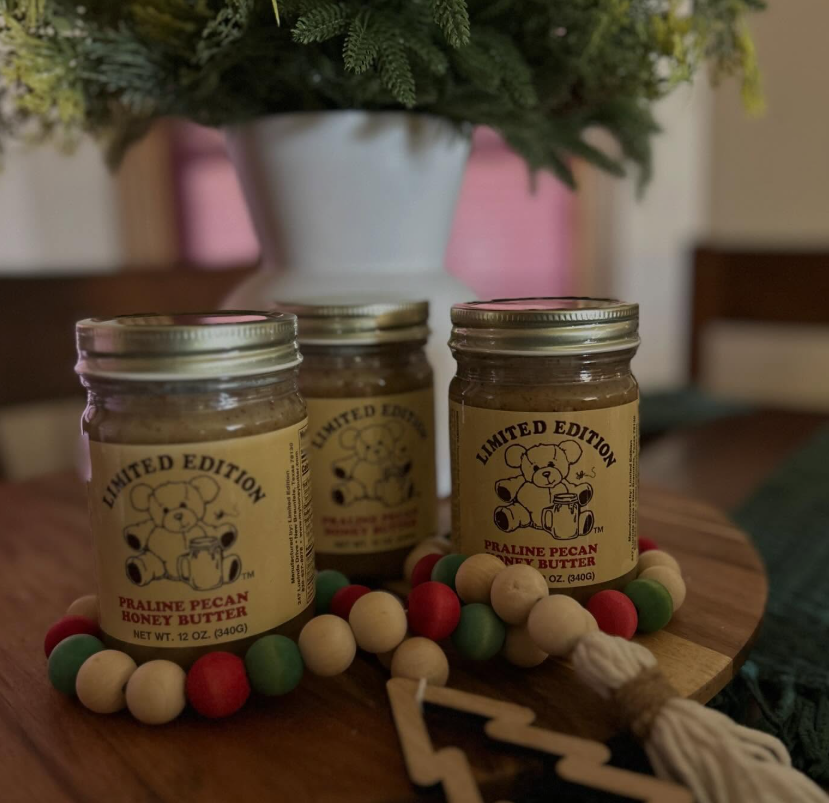 Three mason jars with festive labels on a wooden table, with colorful wooden bead garland and a blurred plant behind.