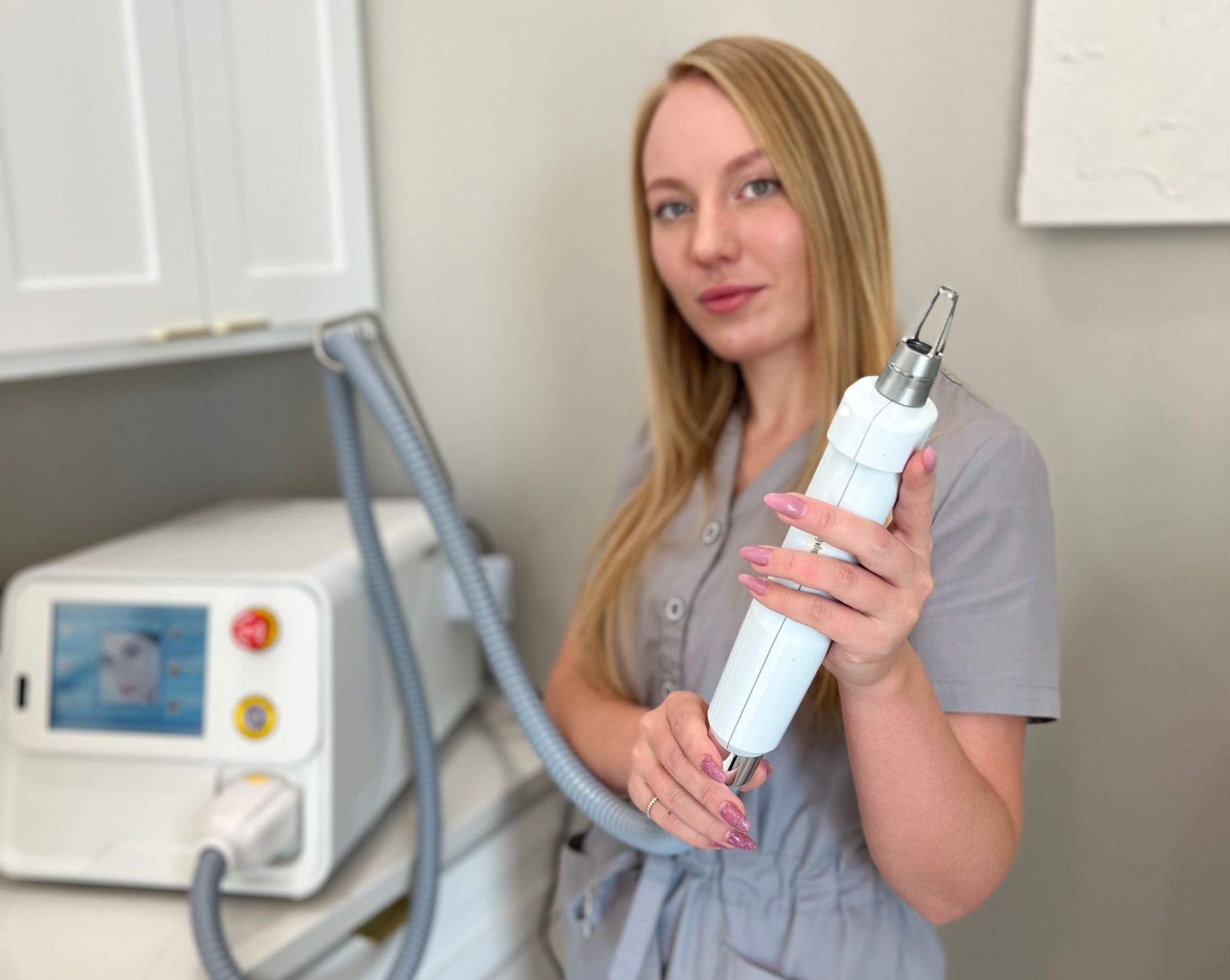 Woman holding a white laser device, standing next to a machine, in a clinic setting.