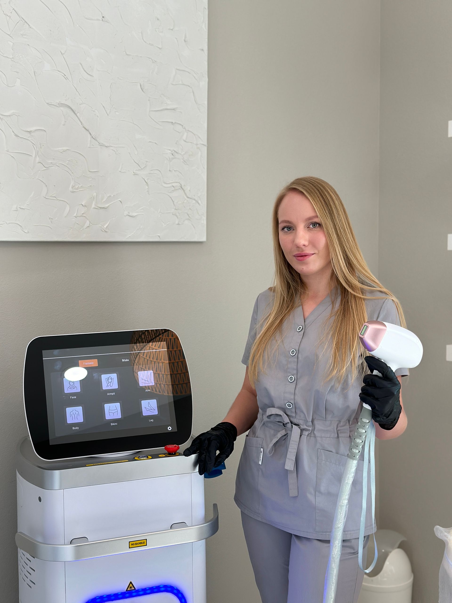 Woman in grey scrubs holding a laser device, standing next to a machine, in a clinic.