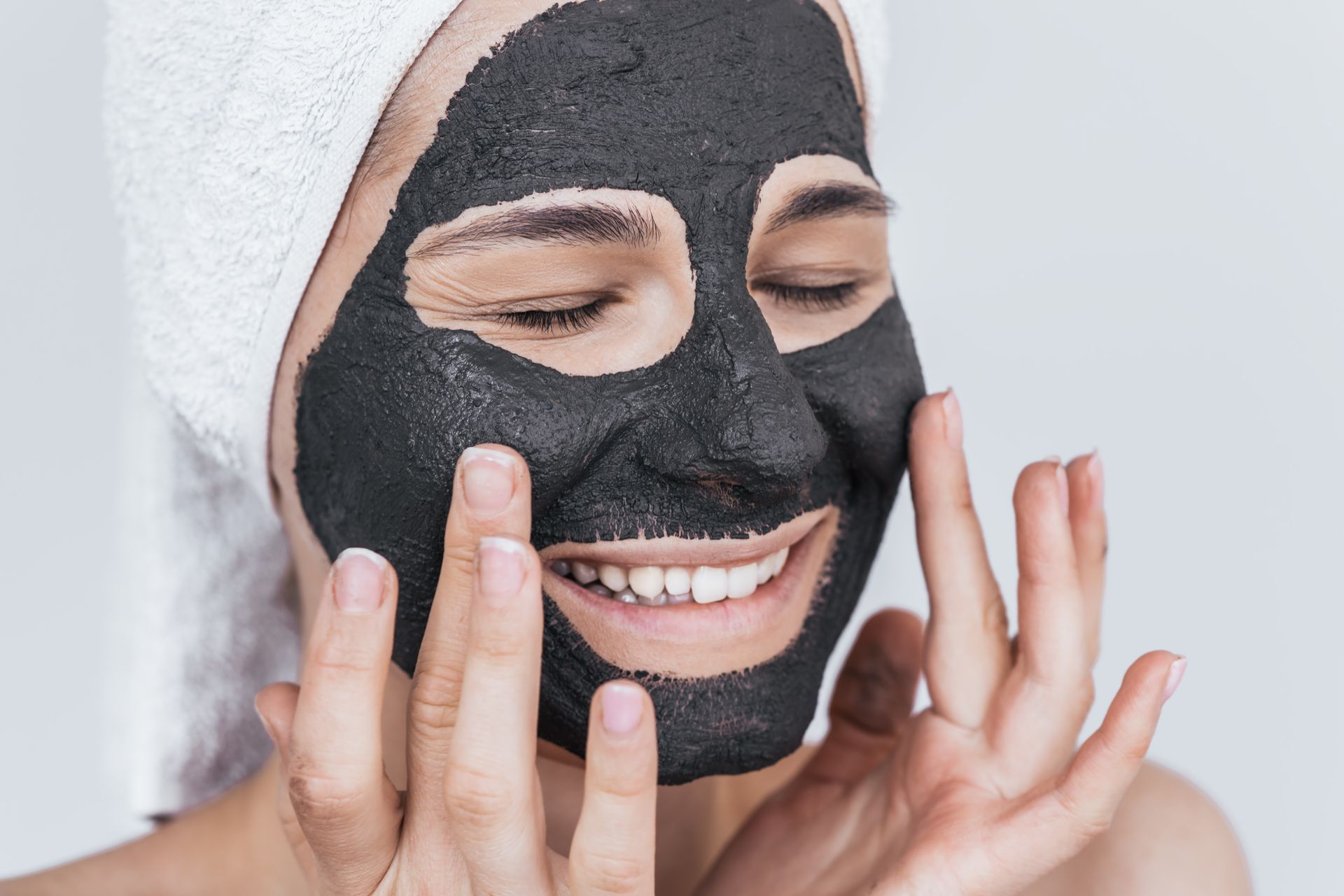 Person with closed eyes and black face mask on, smiling. Hands touching face. White towel on head.