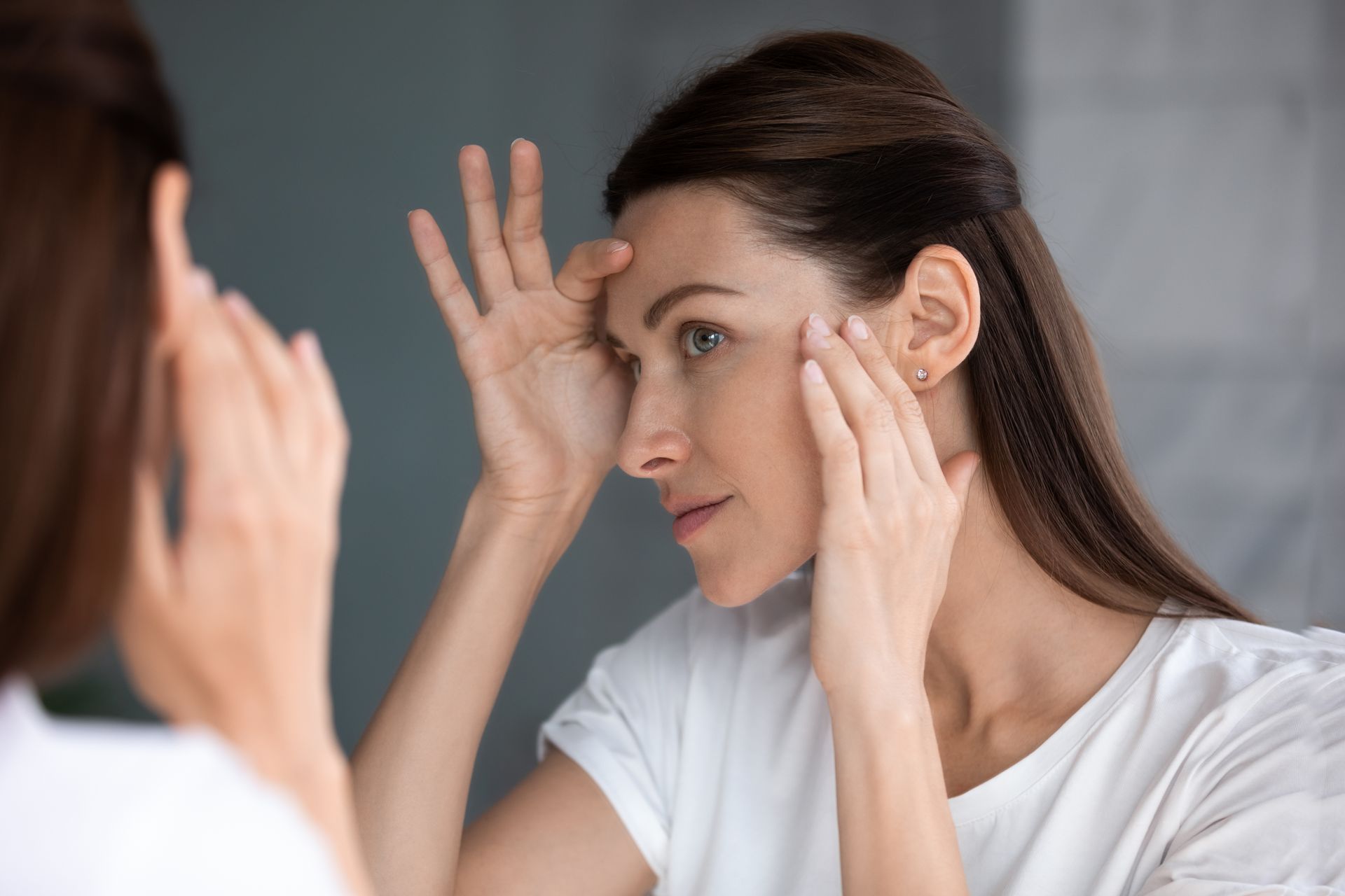 Woman examining her face in a mirror, touching her forehead and cheeks, bathroom setting.