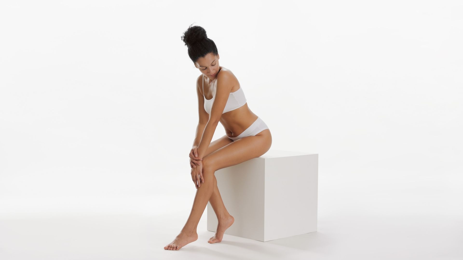 Woman in white lingerie sits on a white cube, touching her leg, against a white background.
