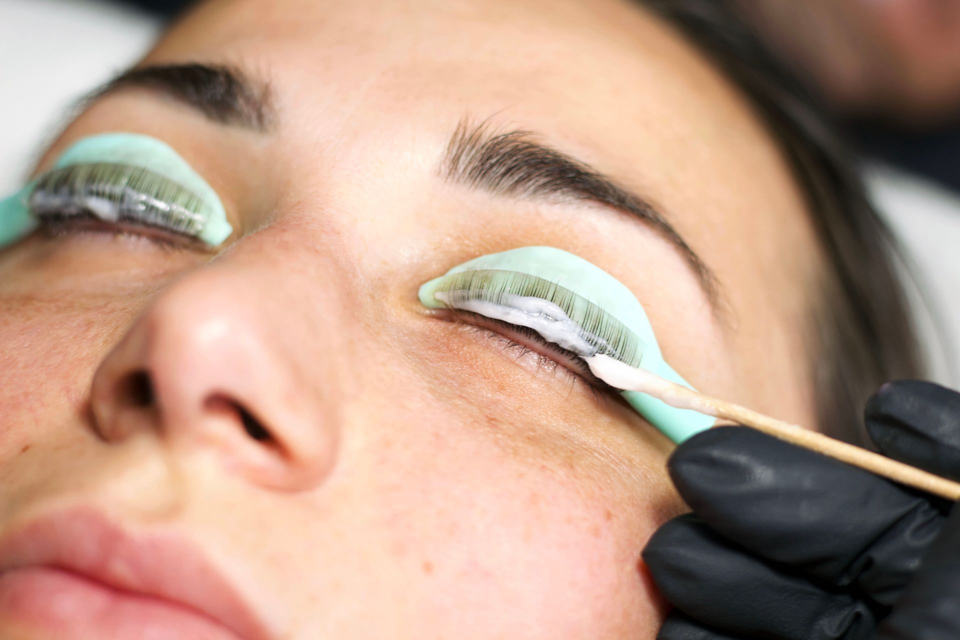 Woman receiving a lash lift treatment with green pads, cotton swab, and black-gloved hands.